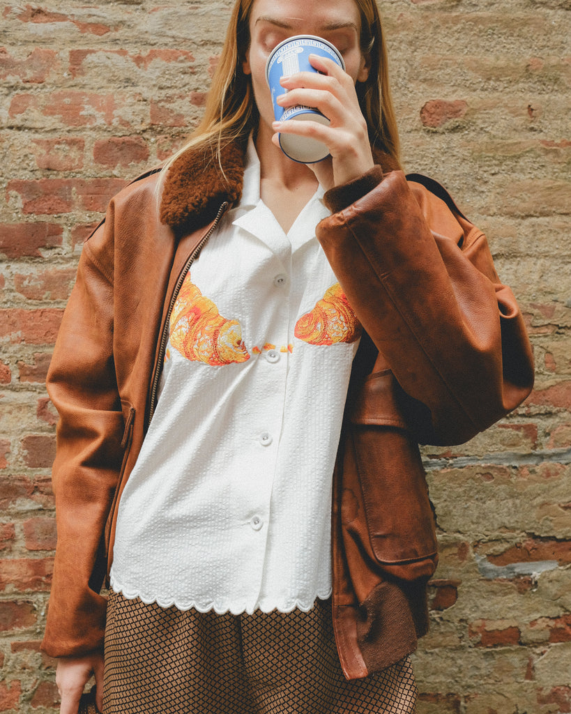 Woman in a white seersucker shirt with croissant embroidery and brown shorts, wearing a brown leather jacket. 