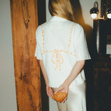 Woman wearing a white shirt with orange bikini embroidery, holding a croissant, in a dimly lit room.
