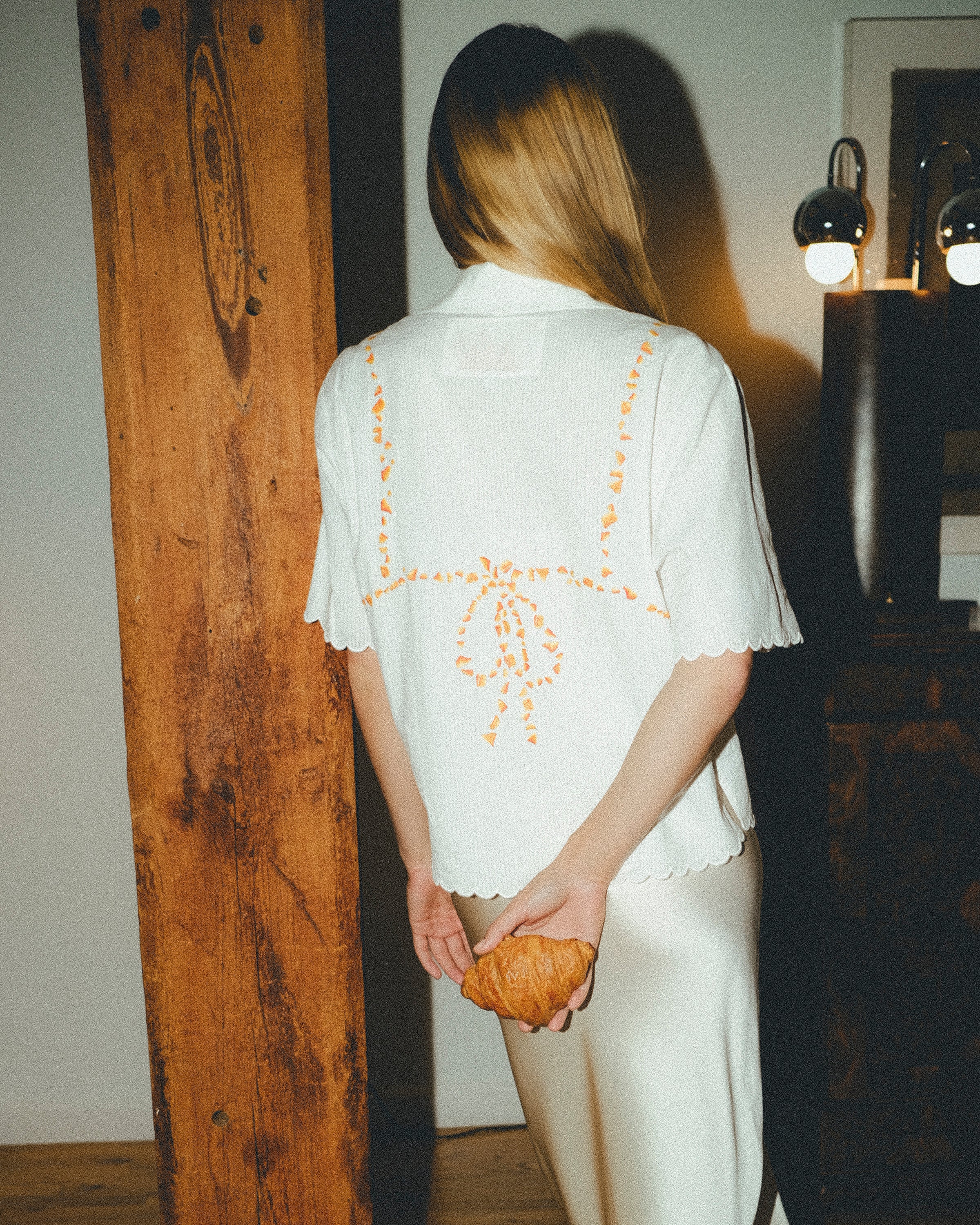 Woman wearing a white shirt with orange bikini embroidery, holding a croissant, in a dimly lit room.