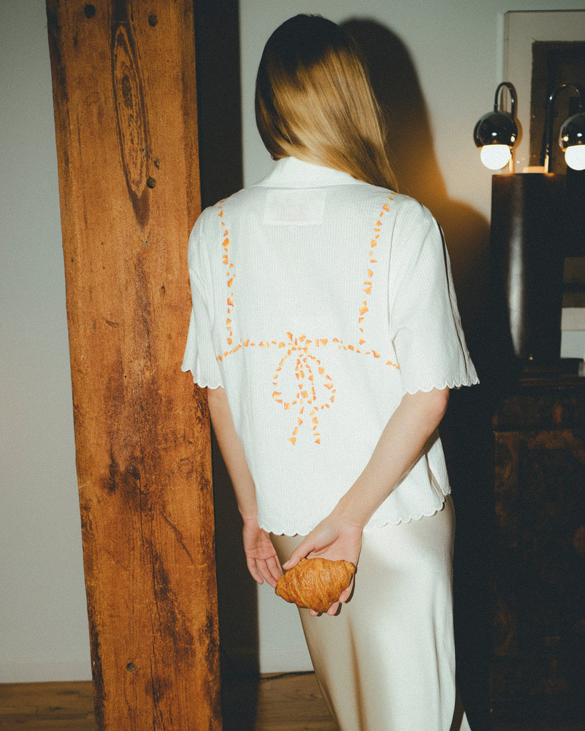 Woman wearing a white shirt with orange bikini embroidery, holding a croissant, in a dimly lit room.