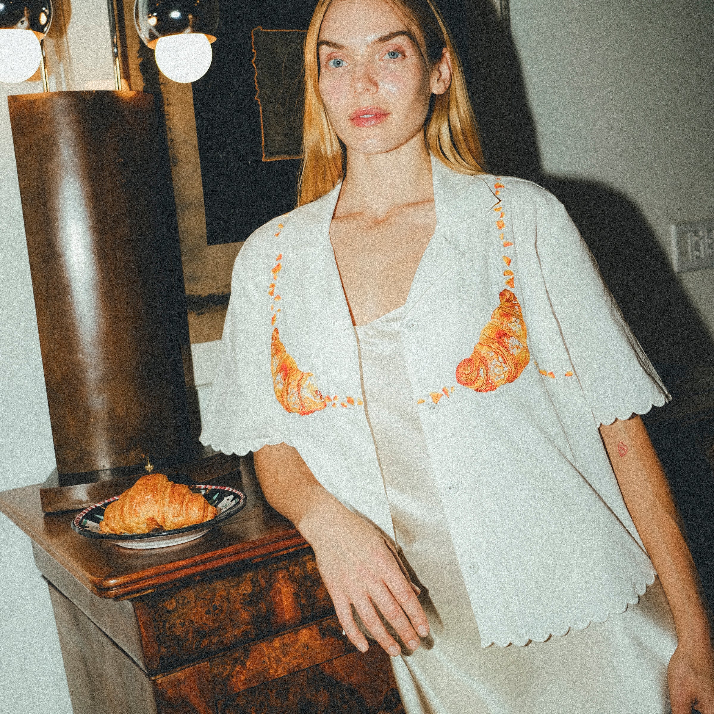 Woman wearing a white embroidered shirt  with a plate of croissants on a wooden table next to her. 