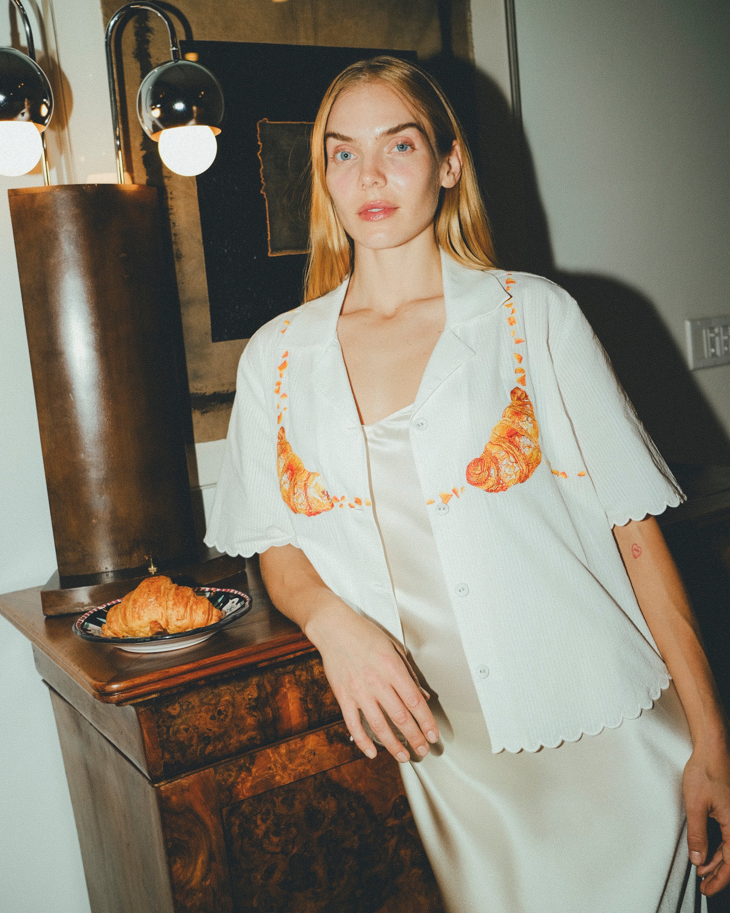 Woman wearing a white embroidered shirt  with a plate of croissants on a wooden table next to her. 