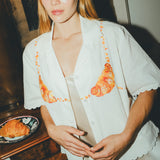Woman wearing a white embroidered shirt  with a plate of croissants on a wooden table next to her. 