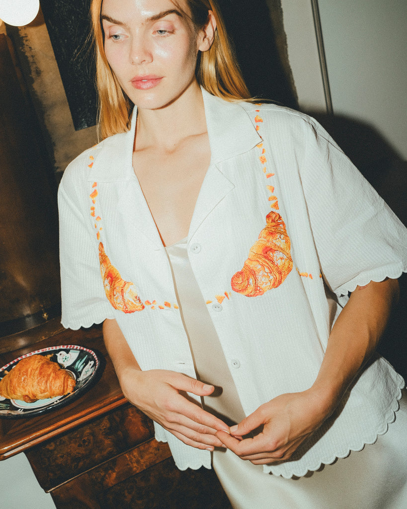 Woman wearing a white embroidered shirt  with a plate of croissants on a wooden table next to her. 
