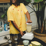 Model wearing a yellow cabana shirt with embroidered cherries and a tropical cocktail, standing at a table while blending a piña colada. Pineapple slices, empty glasses, and liquor bottles are visible in the scene.