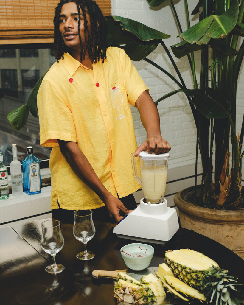 Model wearing a yellow cabana shirt with embroidered cherries and a tropical cocktail, standing at a table while blending a piña colada. Pineapple slices, empty glasses, and liquor bottles are visible in the scene.