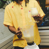 Model wearing a yellow cabana shirt with embroidered cherries and a tropical cocktail design, holding a freshly blended piña colada while pouring from a blender.
