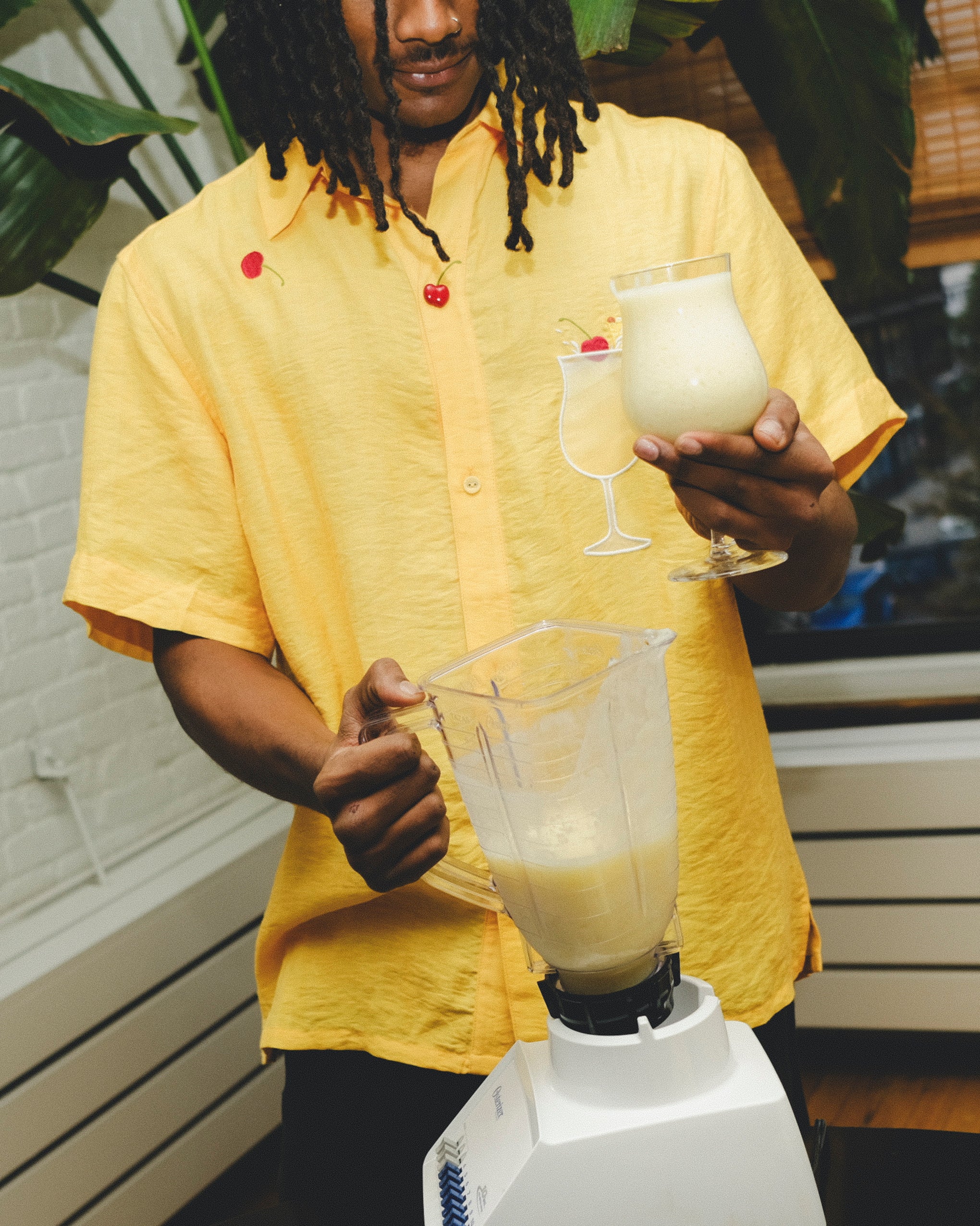 Model wearing a yellow cabana shirt with embroidered cherries and a tropical cocktail design, holding a freshly blended piña colada while pouring from a blender.