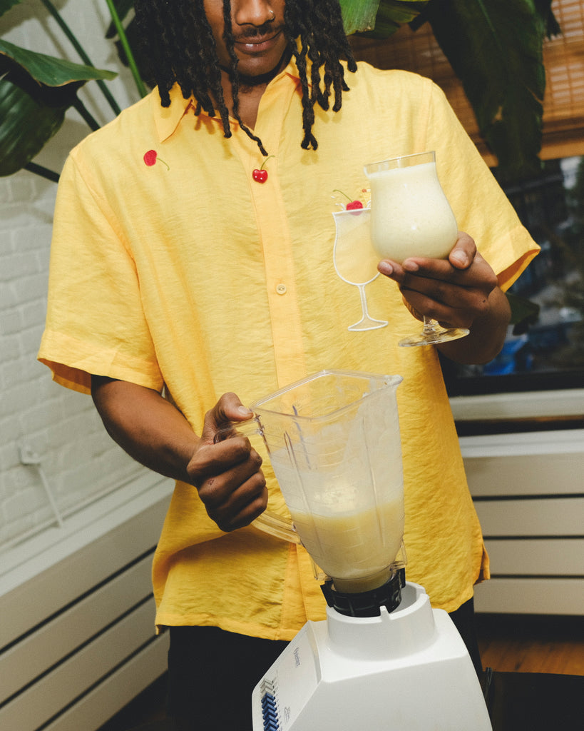 Model wearing a yellow cabana shirt with embroidered cherries and a tropical cocktail design, holding a freshly blended piña colada while pouring from a blender.