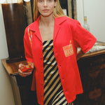 Model wearing a red cabana shirt with twisted appliqué stripes and an embroidered Negroni glass on the chest pocket, styled over a black-and-cream striped dress. The model holds a Negroni-style drink while standing indoors.
