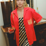 Model wearing a red cabana shirt with twisted appliqué stripes and an embroidered Negroni glass on the chest pocket, styled over a black-and-cream striped dress. The model holds a Negroni-style drink while standing indoors.