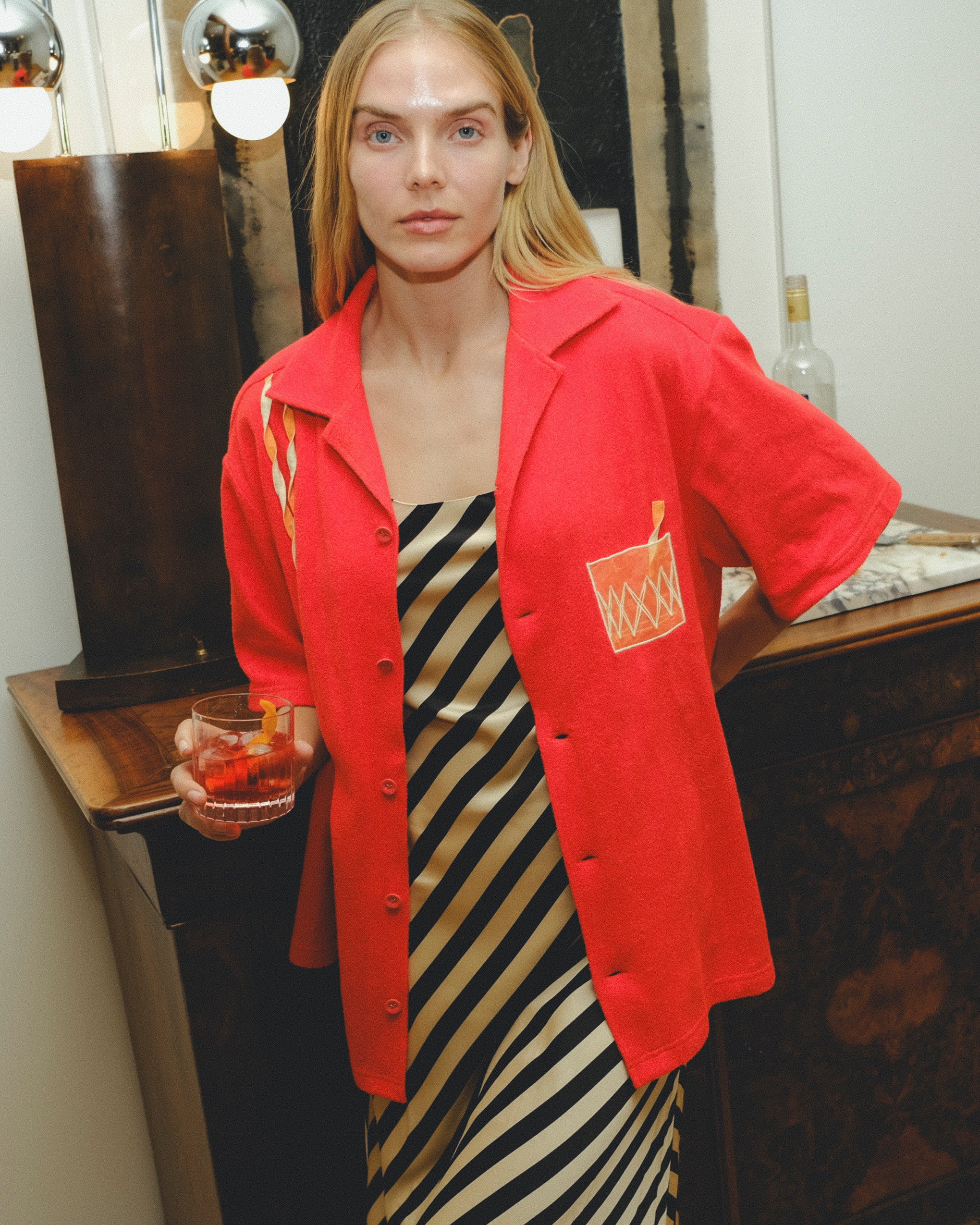 Model wearing a red cabana shirt with twisted appliqué stripes and an embroidered Negroni glass on the chest pocket, styled over a black-and-cream striped dress. The model holds a Negroni-style drink while standing indoors.