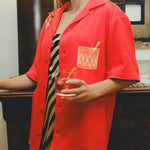 Model wearing a red cabana shirt with an embroidered Negroni glass on the chest pocket, styled over a striped dress. The model holds a Negroni-style drink!