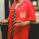 Model wearing a red cabana shirt with an embroidered Negroni glass on the chest pocket, styled over a striped dress. The model holds a Negroni-style drink!