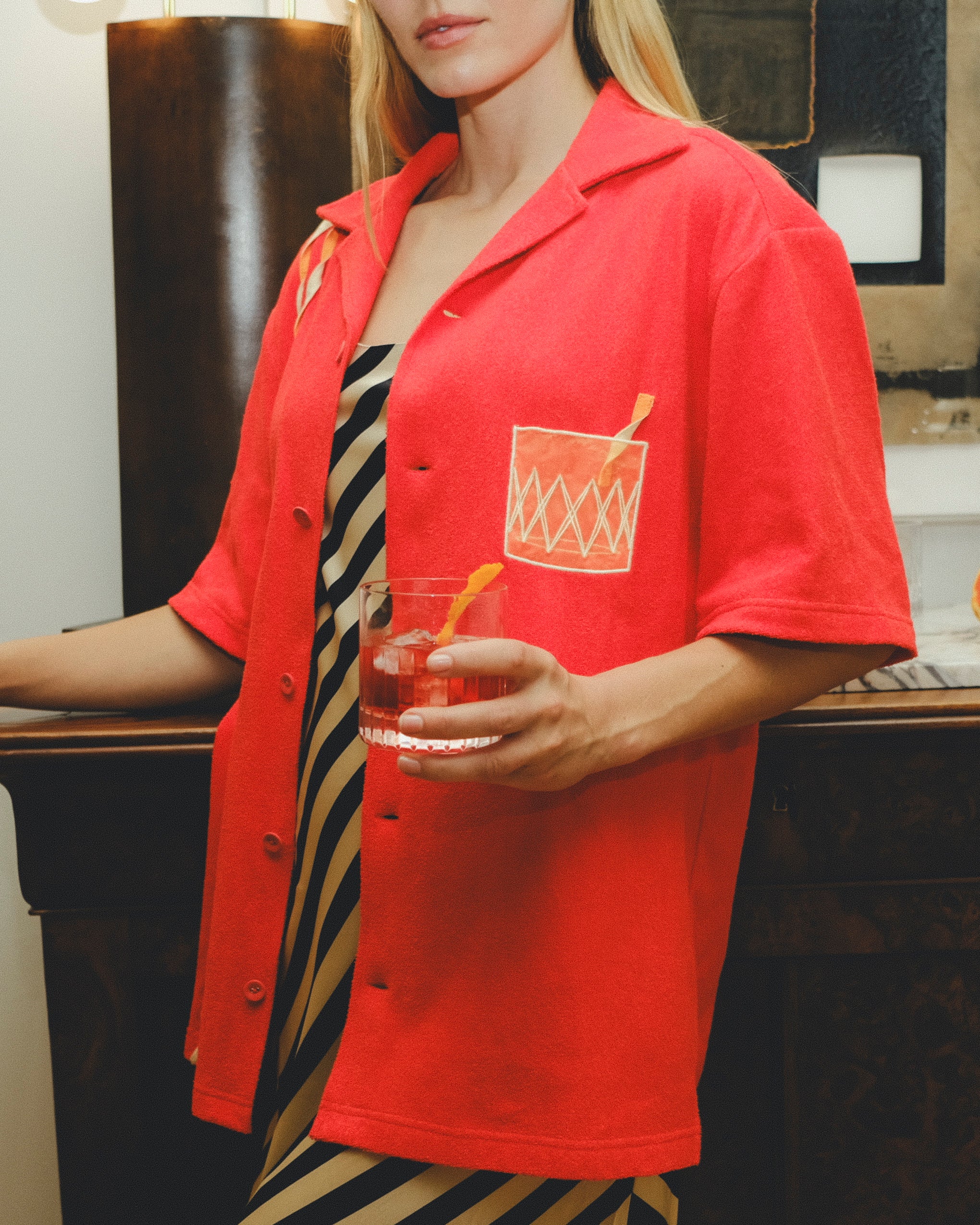 Model wearing a red cabana shirt with an embroidered Negroni glass on the chest pocket, styled over a striped dress. The model holds a Negroni-style drink!