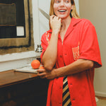Model wearing a red cabana shirt with twisted appliqué stripes and an embroidered Negroni glass on the chest pocket, smiling while holding a kitchen knife and an orange in a food-prep setting.