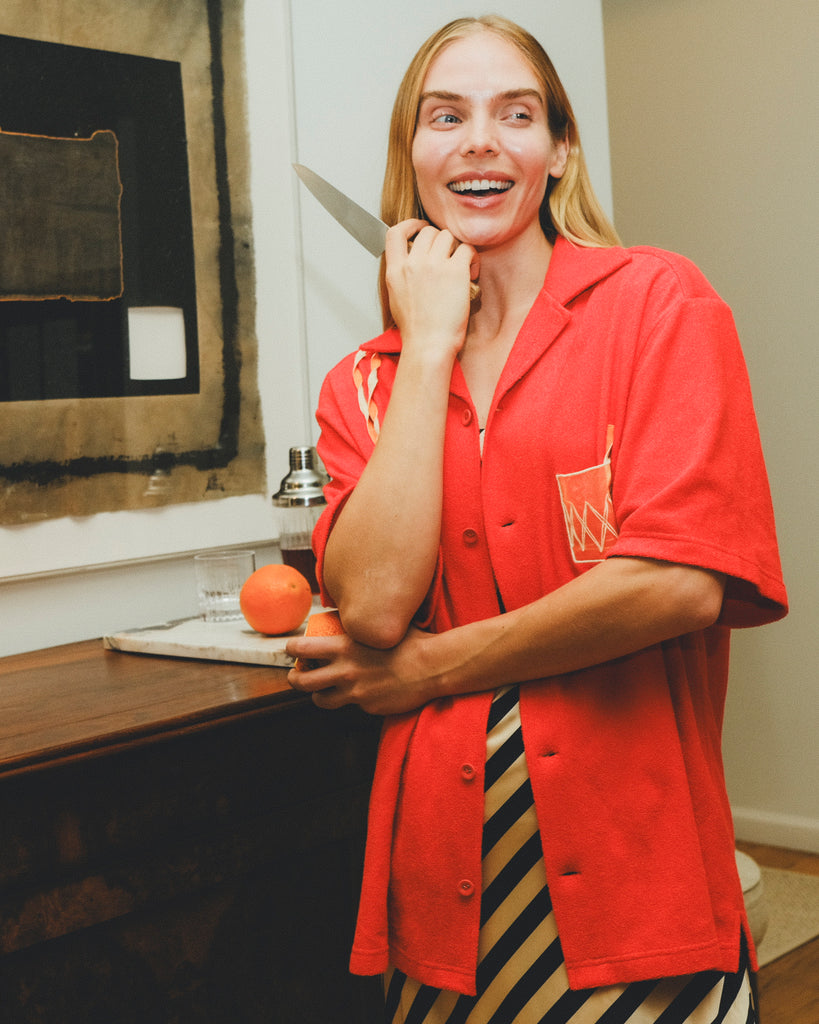 Model wearing a red cabana shirt with twisted appliqué stripes and an embroidered Negroni glass on the chest pocket, smiling while holding a kitchen knife and an orange in a food-prep setting.
