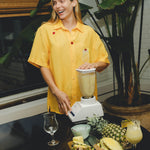 Smiling person wearing a yellow cabana shirt with embroidered cherries and a tropical cocktail, blending a piña colada at a table with pineapples and empty glasses.
