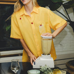 Smiling person wearing a yellow cabana shirt with embroidered cherries and a tropical cocktail, blending a piña colada at a table with pineapples and empty glasses.