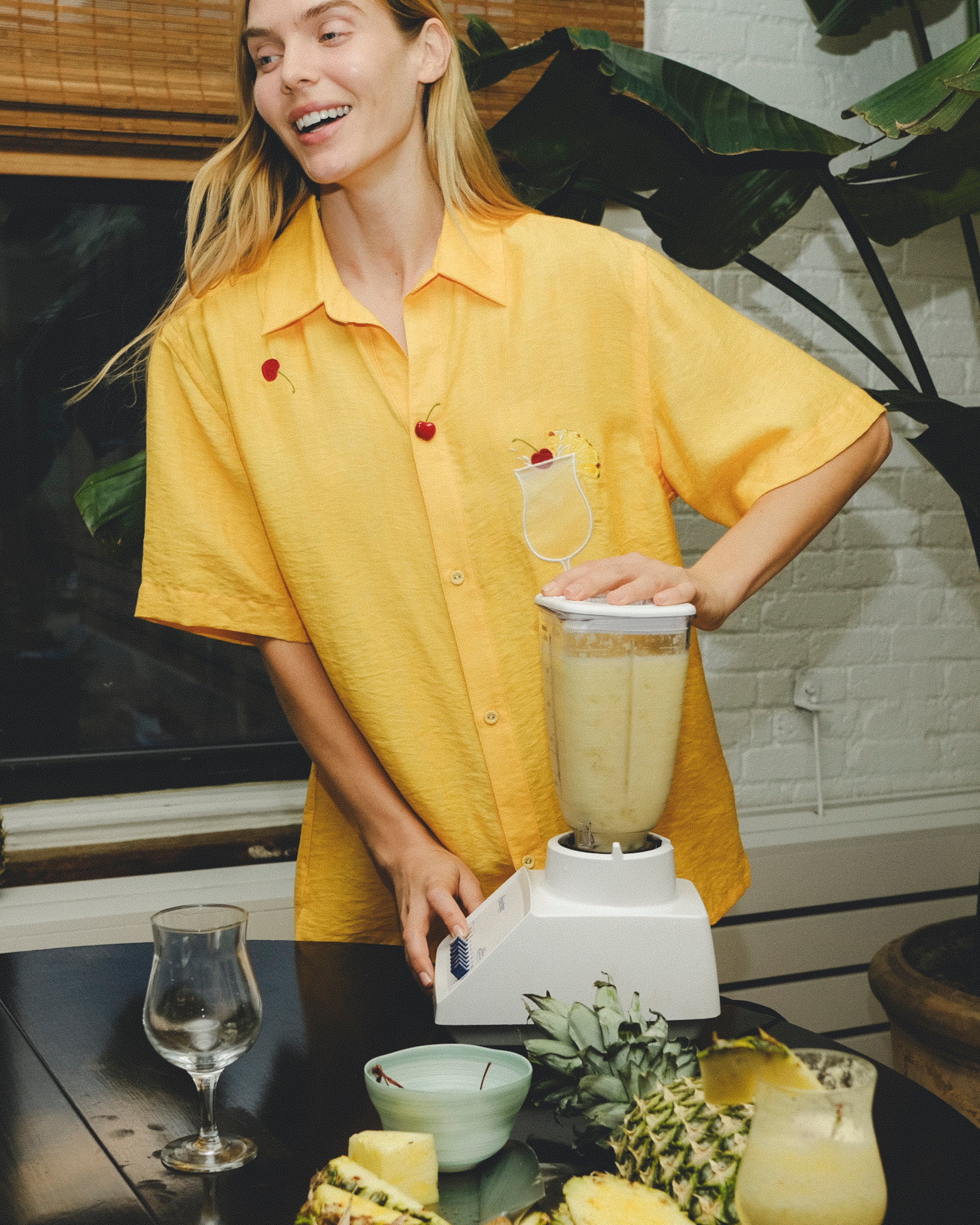 Smiling person wearing a yellow cabana shirt with embroidered cherries and a tropical cocktail, blending a piña colada at a table with pineapples and empty glasses.