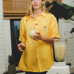 Model wearing a yellow cabana shirt with embroidered cherries and a tropical cocktail, holding a glass of piña colada while standing beside a blender and fresh pineapples.