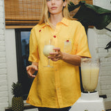 Model wearing a yellow cabana shirt with embroidered cherries and a tropical cocktail, holding a glass of piña colada while standing beside a blender and fresh pineapples.