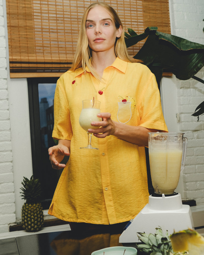 Model wearing a yellow cabana shirt with embroidered cherries and a tropical cocktail, holding a glass of piña colada while standing beside a blender and fresh pineapples.