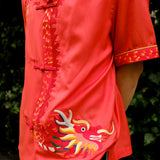 A person wearing a vibrant coral pink short-sleeved shirt with traditional Chinese frog button closures and a mandarin collar, paired with dark striped trousers, showcasing an intricate golden dragon embroidery with multicolored details on the right side. 