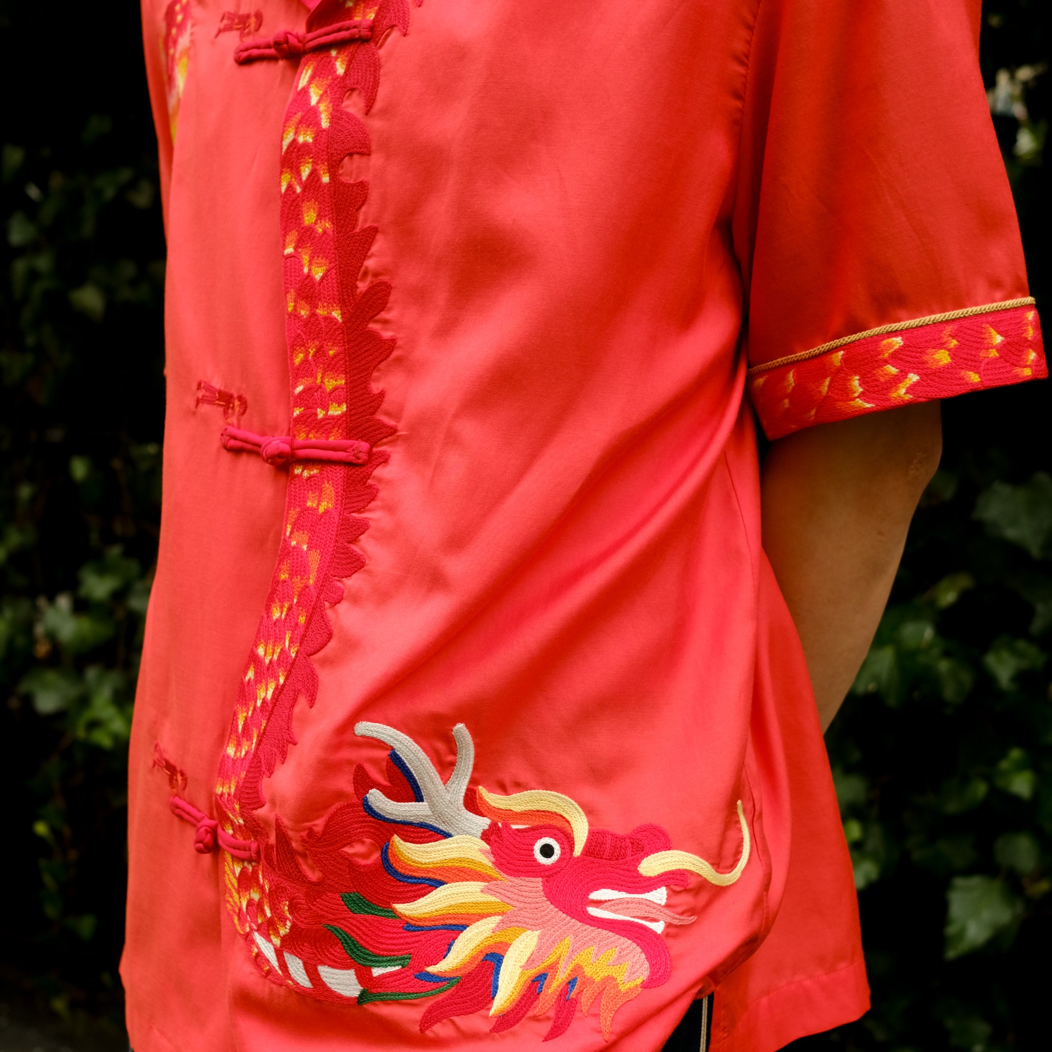 A person wearing a vibrant coral pink short-sleeved shirt with traditional Chinese frog button closures and a mandarin collar, paired with dark striped trousers, showcasing an intricate golden dragon embroidery with multicolored details on the right side. 