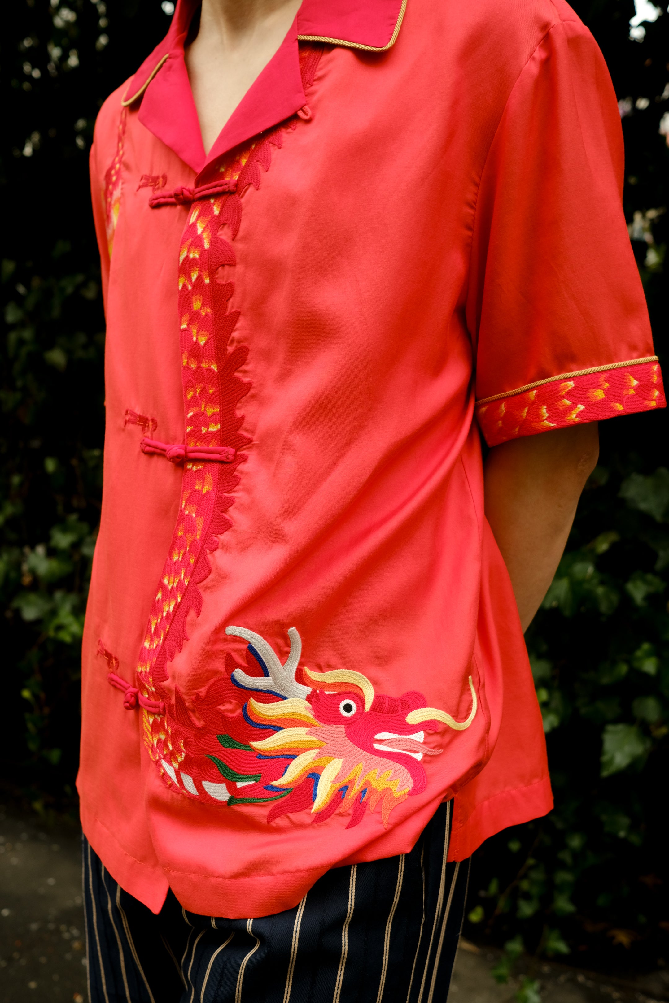 A person wearing a vibrant coral pink short-sleeved shirt with traditional Chinese frog button closures and a mandarin collar, paired with dark striped trousers, showcasing an intricate golden dragon embroidery with multicolored details on the right side. 