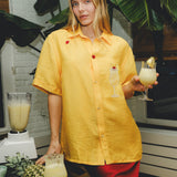 Woman wearing a yellow cabana shirt with embroidered cherries and a tropical cocktail, standing in a kitchen setting with pineapples, a blender, and a piña colada in hand.
