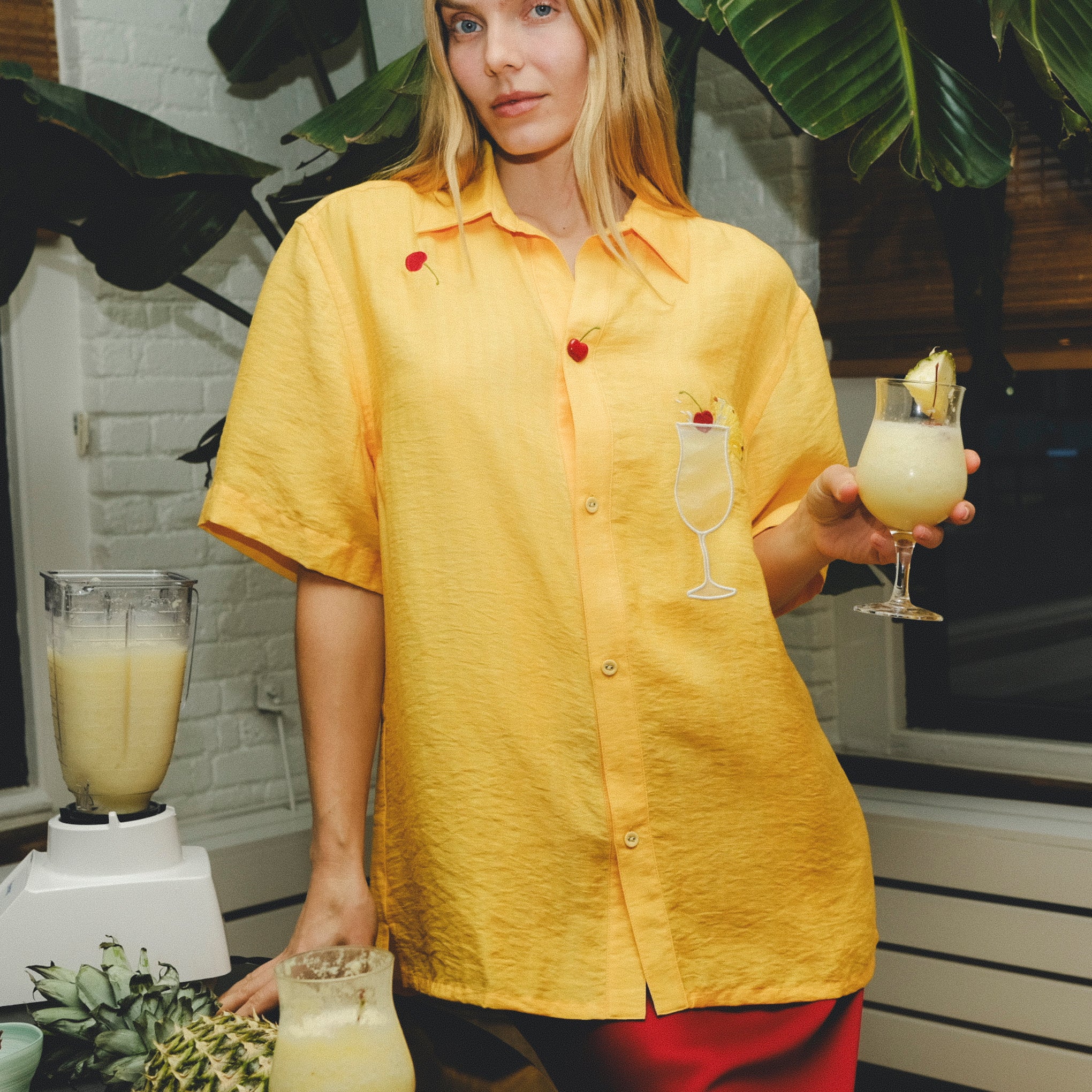 Woman wearing a yellow cabana shirt with embroidered cherries and a tropical cocktail, standing in a kitchen setting with pineapples, a blender, and a piña colada in hand.