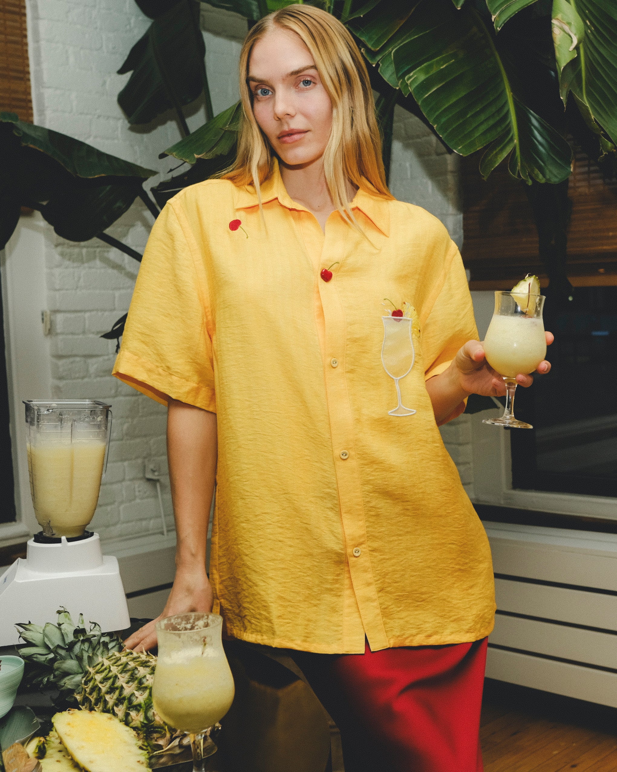 Woman wearing a yellow cabana shirt with embroidered cherries and a tropical cocktail, standing in a kitchen setting with pineapples, a blender, and a piña colada in hand.