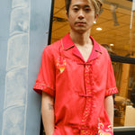 Jiannan Wu stares at the camera with hands in pocket, Tombolo store in the background, displaying his Wood dragon designed red cabana shirt