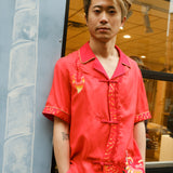 Jiannan Wu stares at the camera with hands in pocket, Tombolo store in the background, displaying his Wood dragon designed red cabana shirt