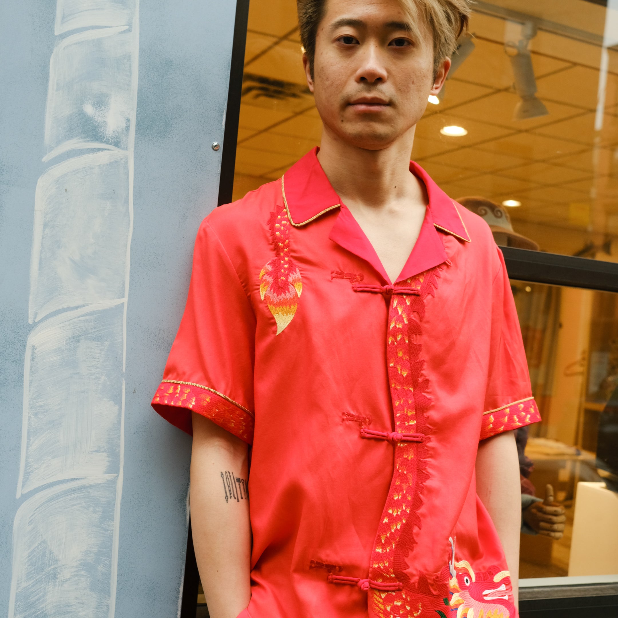 Jiannan Wu stares at the camera with hands in pocket, Tombolo store in the background, displaying his Wood dragon designed red cabana shirt