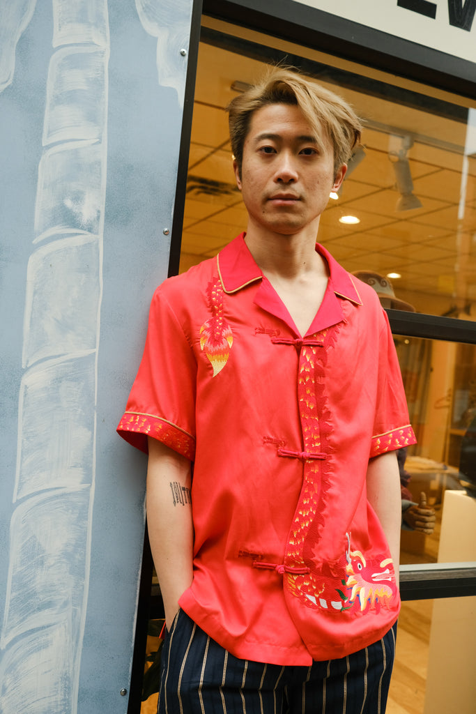 Jiannan Wu stares at the camera with hands in pocket, Tombolo store in the background, displaying his Wood dragon designed red cabana shirt