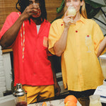 Two models are happily sipping a cocktails while standing indoors with a bar in front of them. The model on the left is male and is wearing a red knit cabana shirt with twisted appliqué stripes and an embroidered Negroni glass on the chest pocket, paired with yellow pants. He is sipping a Negroni-style drink. The model on the right is wearing a yellow woven cabana shirt with embroidered cherries and an embroidered Piña Colada style drink on the chest pocket. She is drinking a Piña Colada style drink.