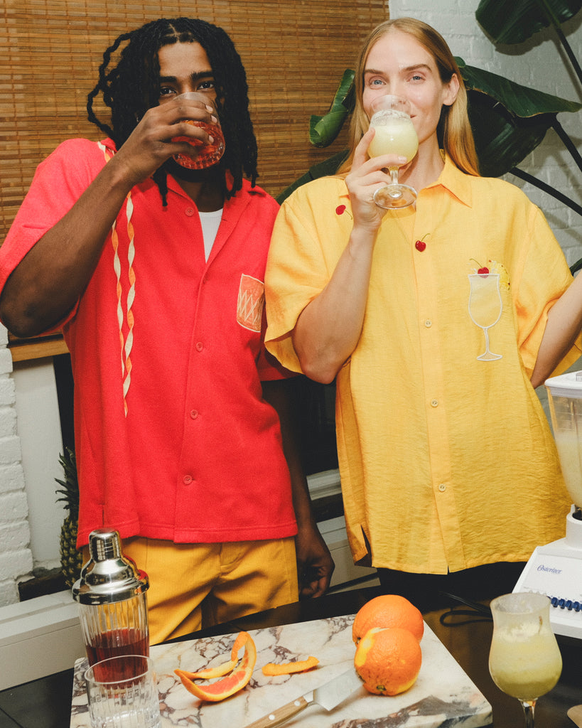Two models are happily sipping a cocktails while standing indoors with a bar in front of them. The model on the left is male and is wearing a red knit cabana shirt with twisted appliqué stripes and an embroidered Negroni glass on the chest pocket, paired with yellow pants. He is sipping a Negroni-style drink. The model on the right is wearing a yellow woven cabana shirt with embroidered cherries and an embroidered Piña Colada style drink on the chest pocket. She is drinking a Piña Colada style drink.