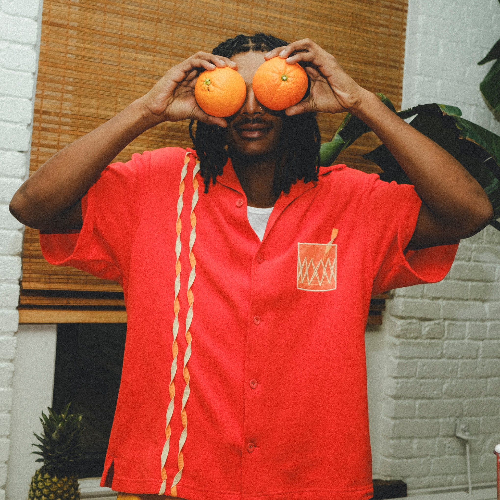 Model wearing a red cabana shirt with twisted appliqué stripes and an embroidered Negroni glass on the chest pocket, holding two oranges over their eyes in a playful pose.