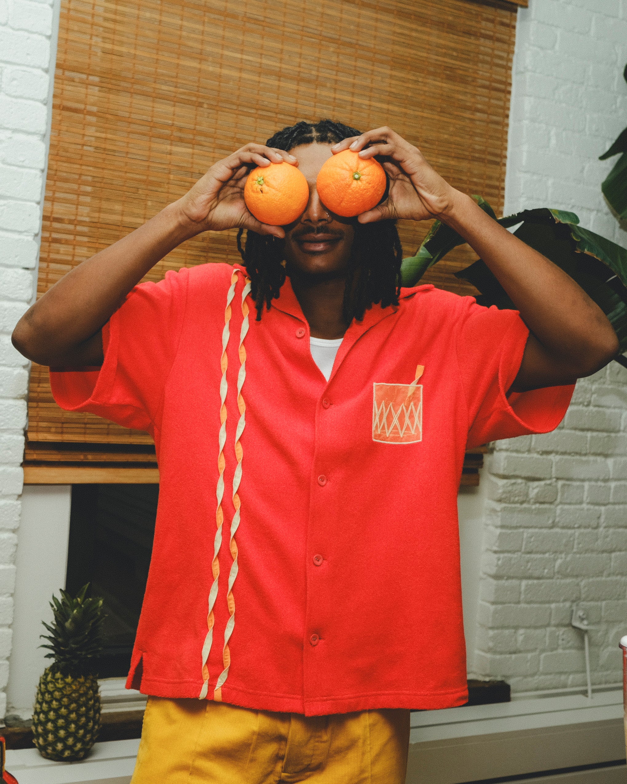 Model wearing a red cabana shirt with twisted appliqué stripes and an embroidered Negroni glass on the chest pocket, holding two oranges over their eyes in a playful pose.
