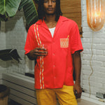 Model wearing a red cabana shirt with twisted appliqué stripes and an embroidered Negroni glass on the chest pocket, paired with yellow pants. The model holds a Negroni-style drink while standing indoors.