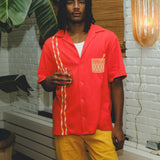 Model wearing a red cabana shirt with twisted appliqué stripes and an embroidered Negroni glass on the chest pocket, paired with yellow pants. The model holds a Negroni-style drink while standing indoors.