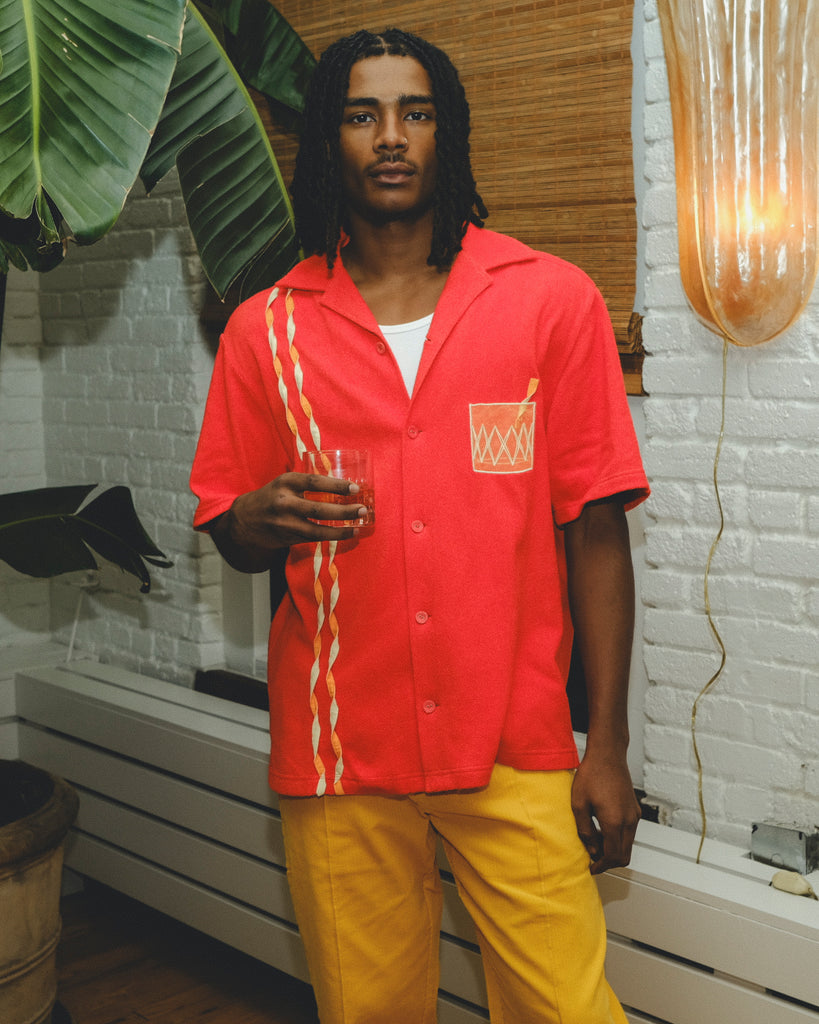 Model wearing a red cabana shirt with twisted appliqué stripes and an embroidered Negroni glass on the chest pocket, paired with yellow pants. The model holds a Negroni-style drink while standing indoors.