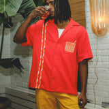 Model wearing a red cabana shirt with twisted appliqué stripes and an embroidered Negroni glass on the chest pocket, paired with yellow pants. The model is sipping a Negroni-style drink while standing indoors.