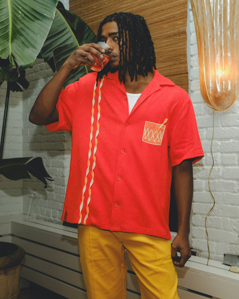 Model wearing a red cabana shirt with twisted appliqué stripes and an embroidered Negroni glass on the chest pocket, paired with yellow pants. The model is sipping a Negroni-style drink while standing indoors.