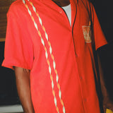 Model wearing a red cabana shirt featuring three twisted appliqué stripes down one side and an embroidered Negroni glass on the chest pocket. 