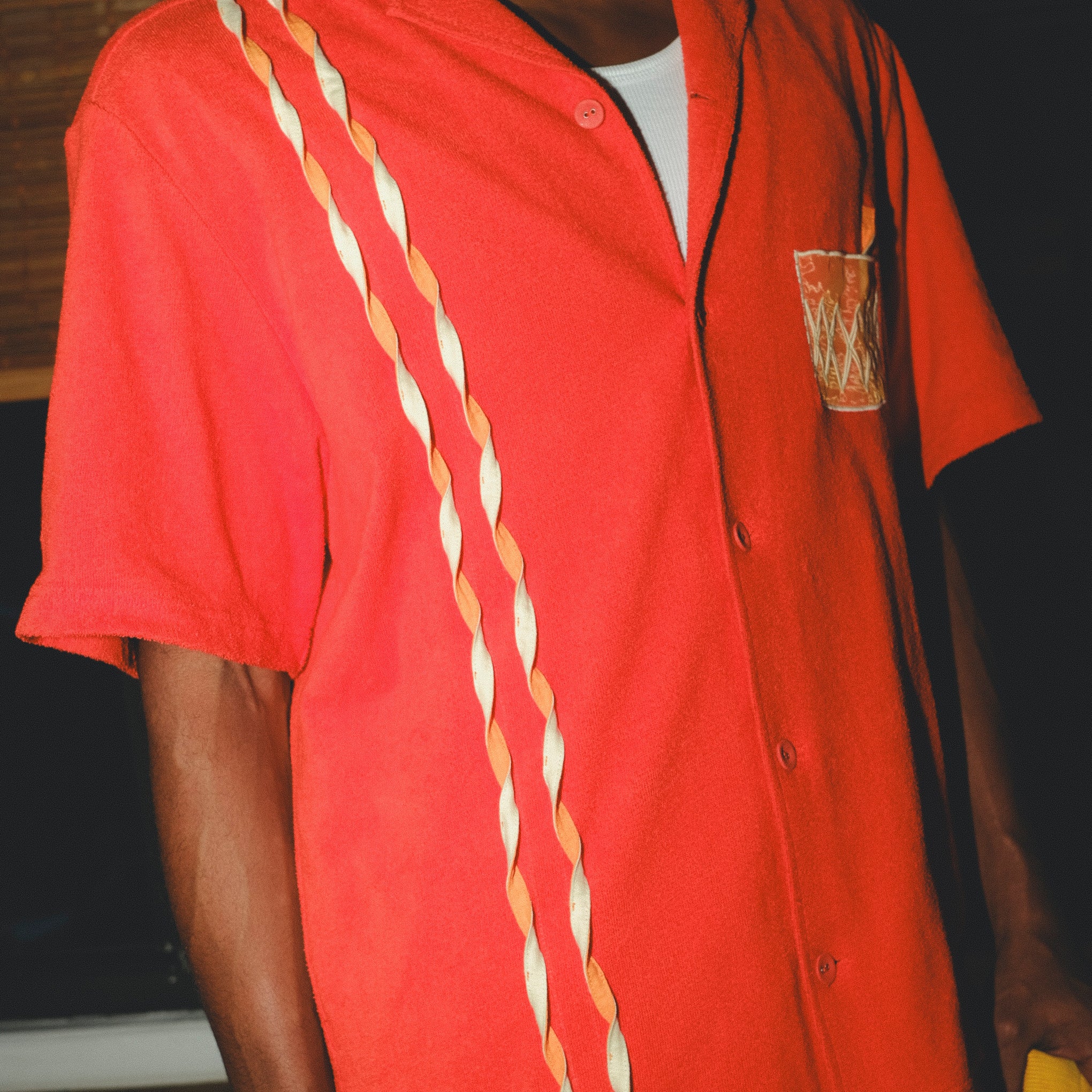 Model wearing a red cabana shirt featuring three twisted appliqué stripes down one side and an embroidered Negroni glass on the chest pocket. 