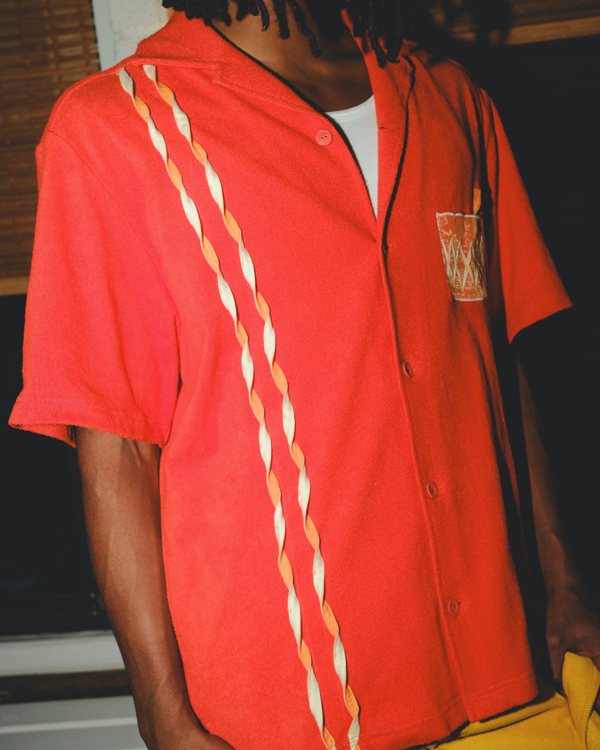 Model wearing a red cabana shirt featuring three twisted appliqué stripes down one side and an embroidered Negroni glass on the chest pocket. 
