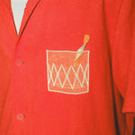 Close-up of a red cabana shirt showing the embroidered Negroni glass on the chest pocket, featuring a transparent mesh panel and stitched orange garnish detail.